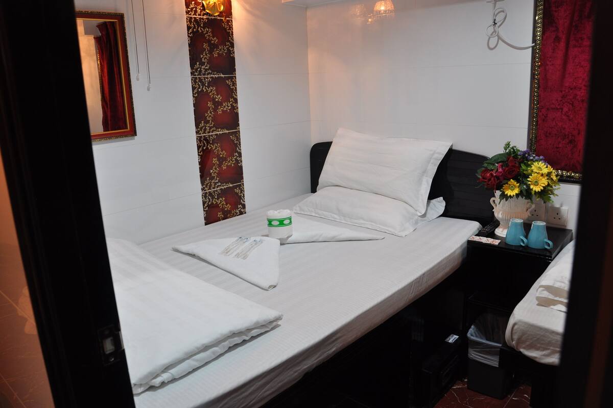 A small, neatly made bed with white linens, a bedside table with a vase of flowers, and a black nightstand with a trash bin.