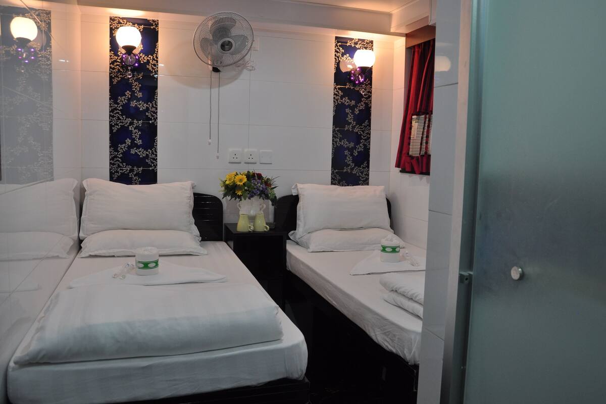A hotel room with two single beds, a bedside table with a vase of flowers, a wall-mounted fan, and decorative wall lights.