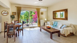 Ein Wohnzimmer mit Essbereich, einer Couch, einem Couchtisch und Blick auf einen Balkon mit Pflanzen und einer rosa Blume.