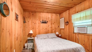 A wooden-paneled room with a bed, bedside lamps, a small nightstand, a mirror, and a window with blinds. - The Nest at Hiding Place Cabins (Logan)
