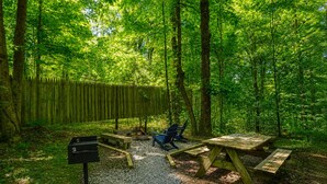 A forested campsite with picnic tables, benches, and a fire pit area. - The Nest at Hiding Place Cabins (Logan)