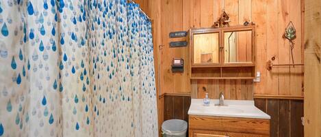 Une salle de bain en bois avec un lavabo, un miroir et une poubelle. Des panneaux indiquent l’emplacement de la salle de bain, et un rideau de douche est orné d’un motif représentant des gouttes d’eau.