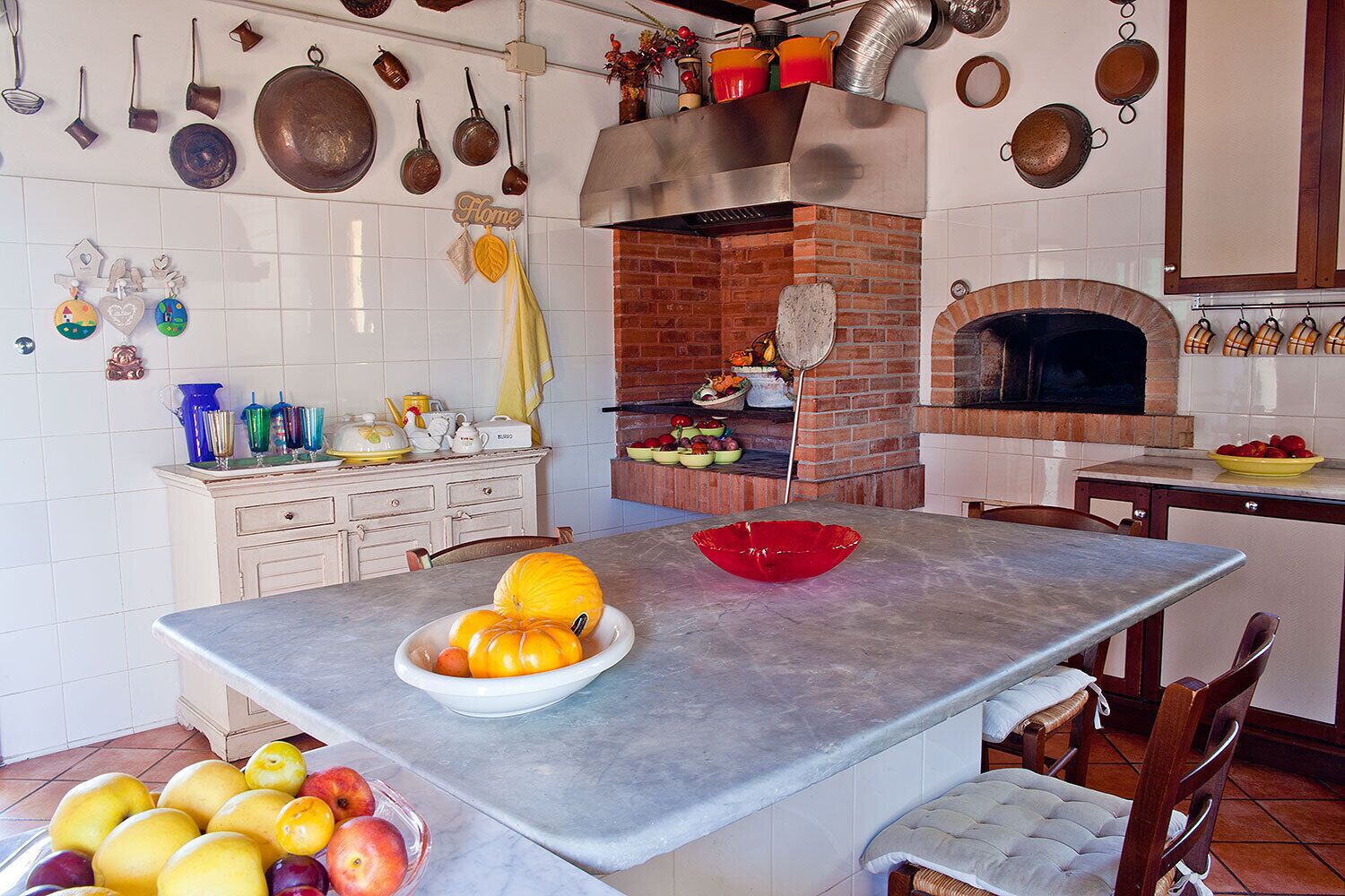 A kitchen with a central island, a brick oven, and various kitchen utensils and decorations on the walls.