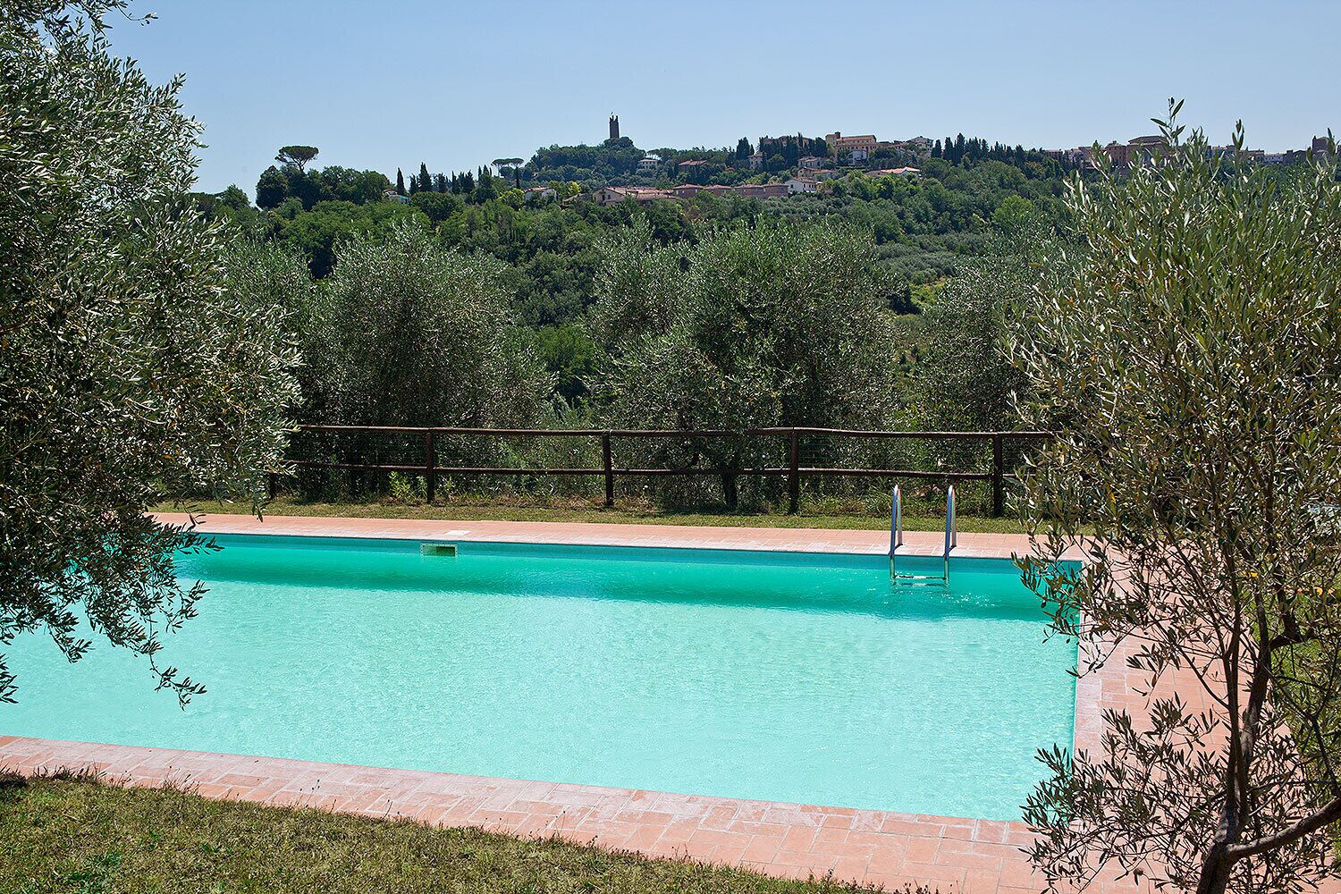 Una piscina rectangular con agua cristalina azul, rodeada por un deck de ladrillo y barandillas metálicas. Al fondo, se divisa una colina con edificios y árboles.