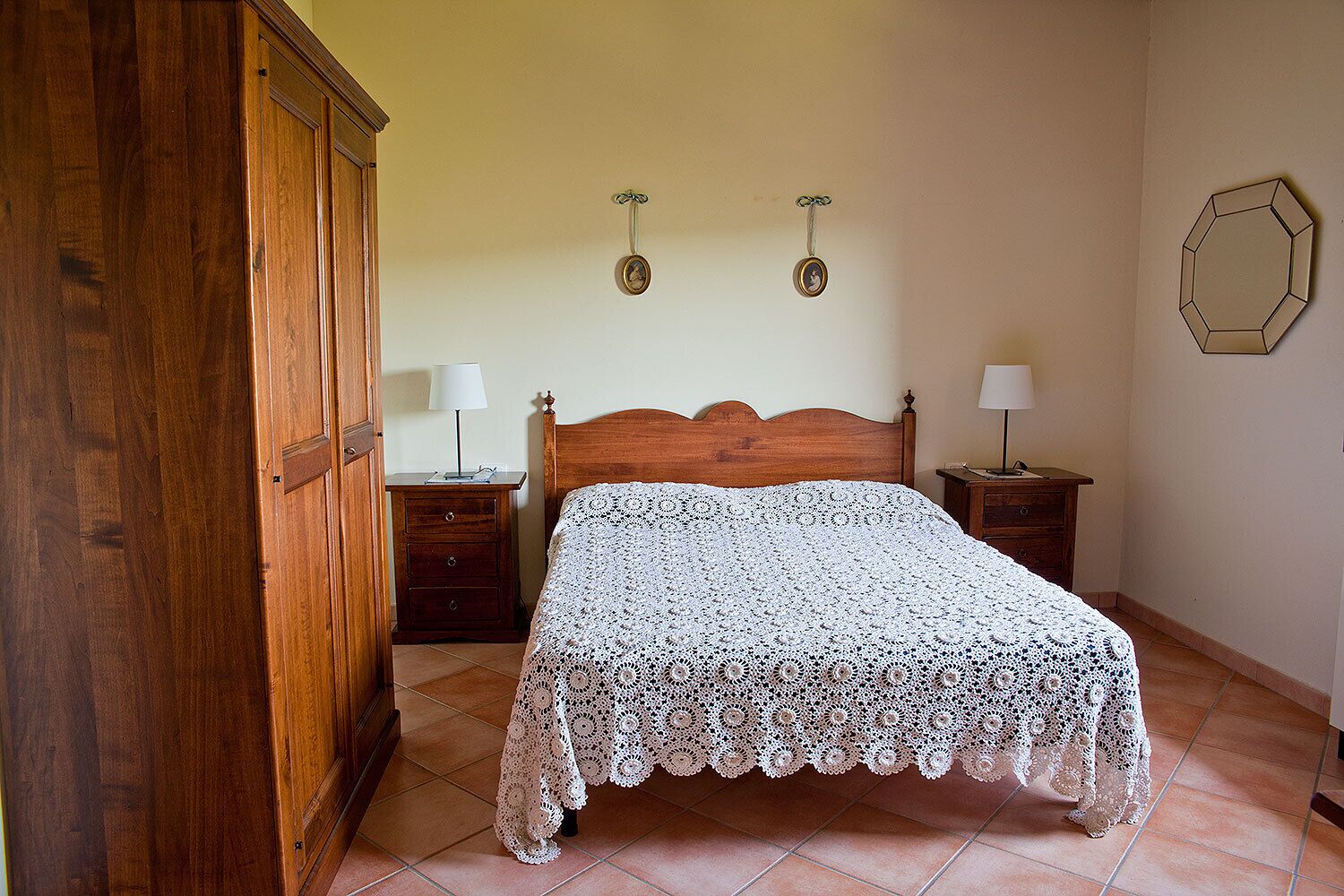 A bedroom with a wooden bed, two bedside tables with lamps, a wooden wardrobe, and a wall clock.