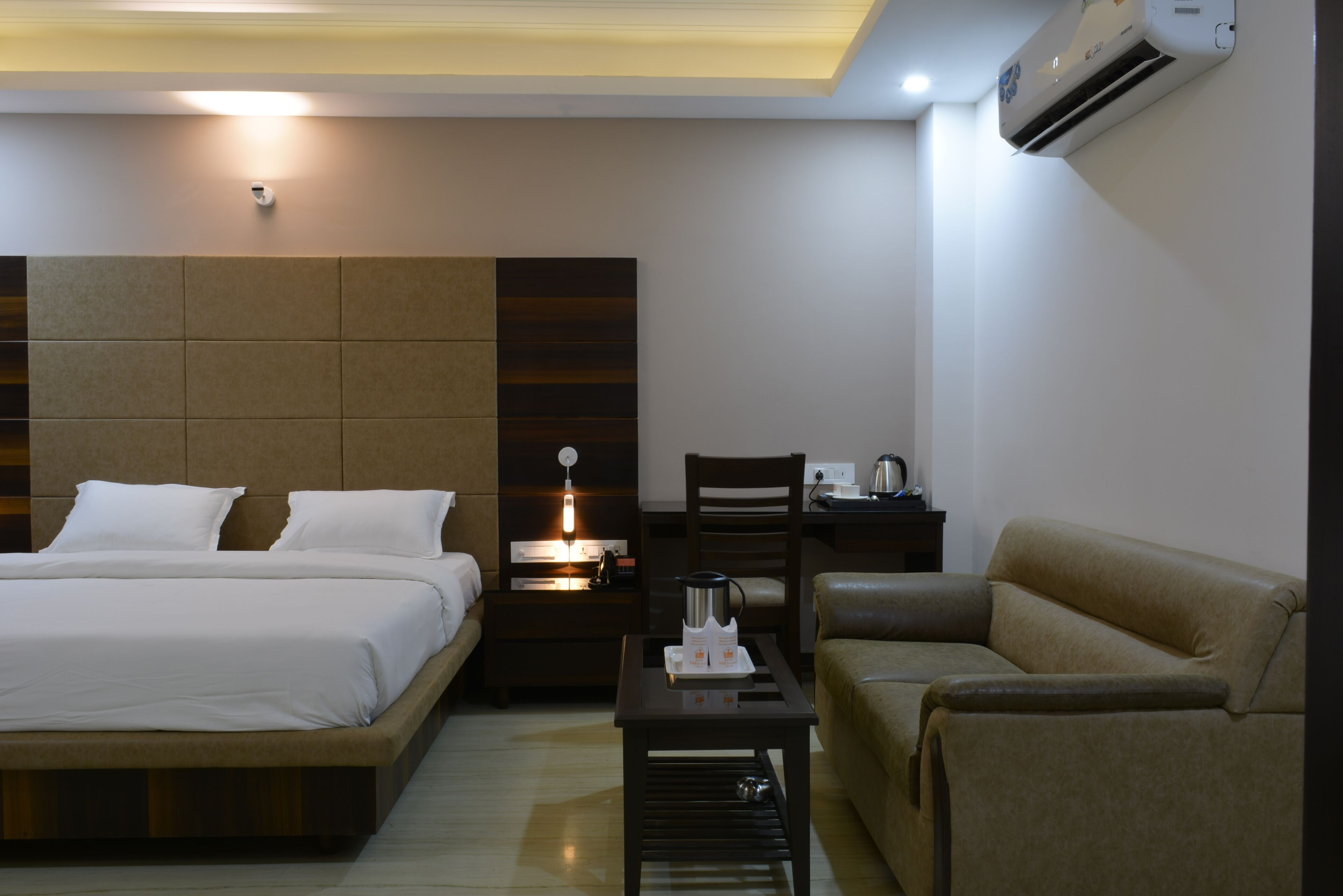 A hotel room with a bed, a nightstand, a coffee table, a chair, and an air conditioning unit.