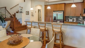 A modern kitchen with wooden cabinets, a stainless steel refrigerator, and a granite countertop. There is a dining area with a glass table and wicker chairs. - Private and Quiet, Luxury Family-Friendly unit in Fairways at Mauna Lani (Waimea)