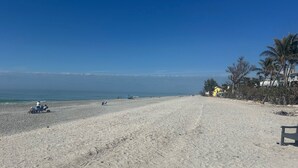 A wide sandy beach with a few people and beach chairs, a clear blue sky, and a building with a yellow facade on the right. - Captiva home- steps from the beach, pool, restaurants and boat dock (Captiva)