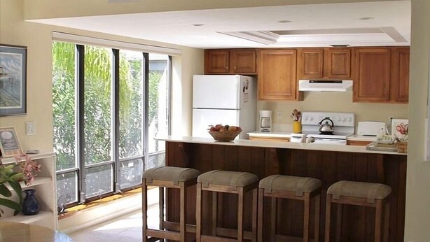 A kitchen with wooden cabinets, a refrigerator, a coffee maker, and a window with a view of greenery. - Captiva home- steps from the beach, pool, restaurants and boat dock (Captiva)
