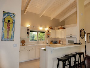 A kitchen with white cabinetry, a central island with stools, a window with a view of greenery, and a wall with a poster.
