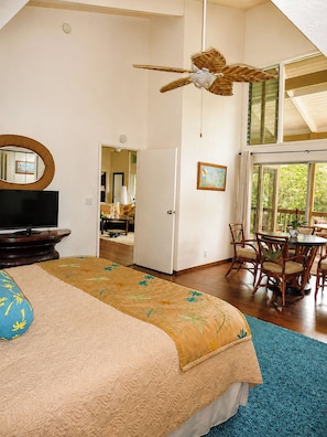 A bedroom with a bed, a television, a ceiling fan, a dining area with chairs and a table, and a window with a view.