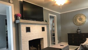 A living room with a fireplace, a flat-screen TV mounted above it, a dark wooden cabinet, and a decorative wall piece. - Walk to Downtown/The Landing! Beautiful cottage, cozy private hot tub! (Branson)