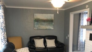 A living room with a black leather sofa, a grey ottoman, and a black armchair with patterned cushions. A ceiling fan is visible above. - Walk to Downtown/The Landing! Beautiful cottage, cozy private hot tub! (Branson)