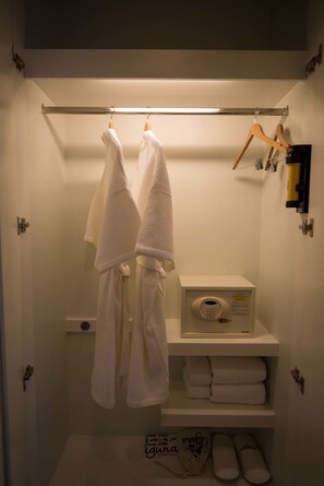 Un baño con un perchero que sostiene dos toallas blancas, un armario blanco con artículos de aseo y una estantería con toallas dobladas y zapatillas.