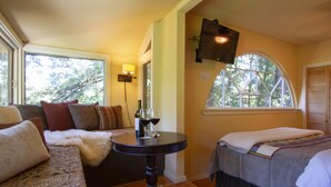 Una habitación acogedora con sofá, cama, una mesita con copas de vino, lámpara y un ventanal con vistas a árboles.