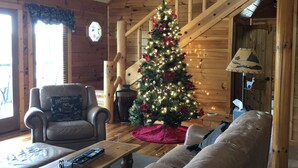 Un acogedor interior de cabaña de madera con una escalera de madera, un árbol de Navidad, sillones de cuero y una chimenea.