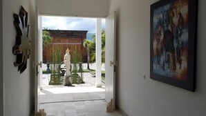 A hallway with a view of an outdoor area featuring a statue, a wooden fence, and a building with a tiled roof. - Villa Cymopoleia (Seririt)