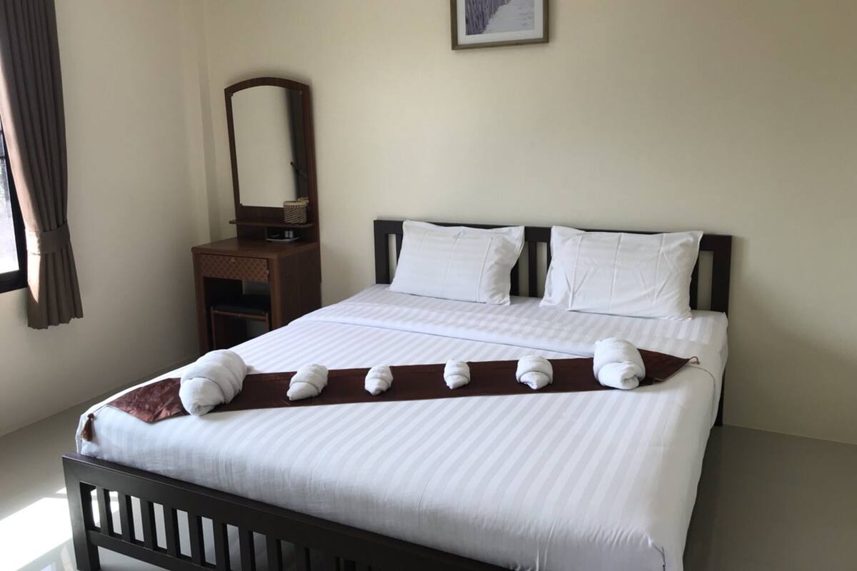 A neatly made bed with white linens and a brown throw blanket, a wooden nightstand with a mirror and a basket, and a framed picture on the wall.