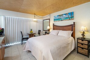 Quarto de hotel com uma cama grande, uma escrivaninha com cadeira, uma mesinha de centro com uma travessa de frutas, uma luminária e uma pintura de uma cena de praia.