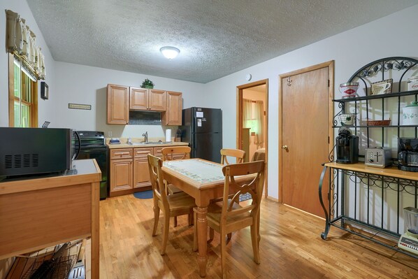 A compact kitchen with wooden cabinets, a dining table with chairs, a refrigerator, and a coffee machine.
