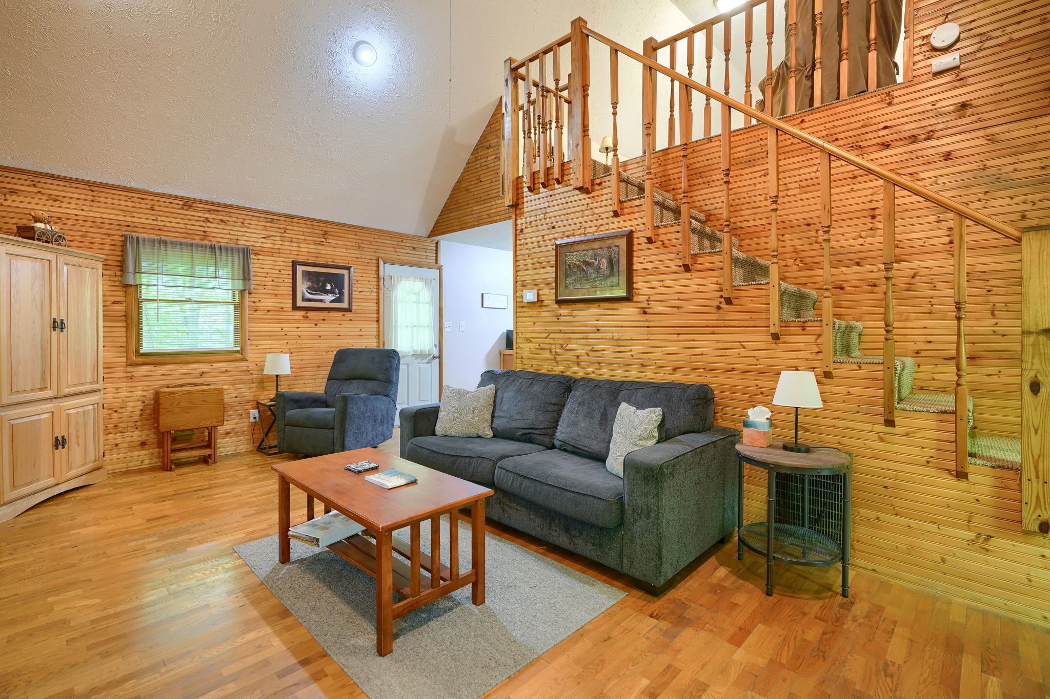 Un salon chaleureux avec un escalier en bois, un canapé gris, une table en bois et une petite table d’appoint sur laquelle se trouve une lampe.