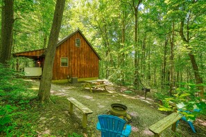 Uma cabana rústica numa floresta, com uma mesa de piquenique, uma cadeira azul e um espaço para fogueira.