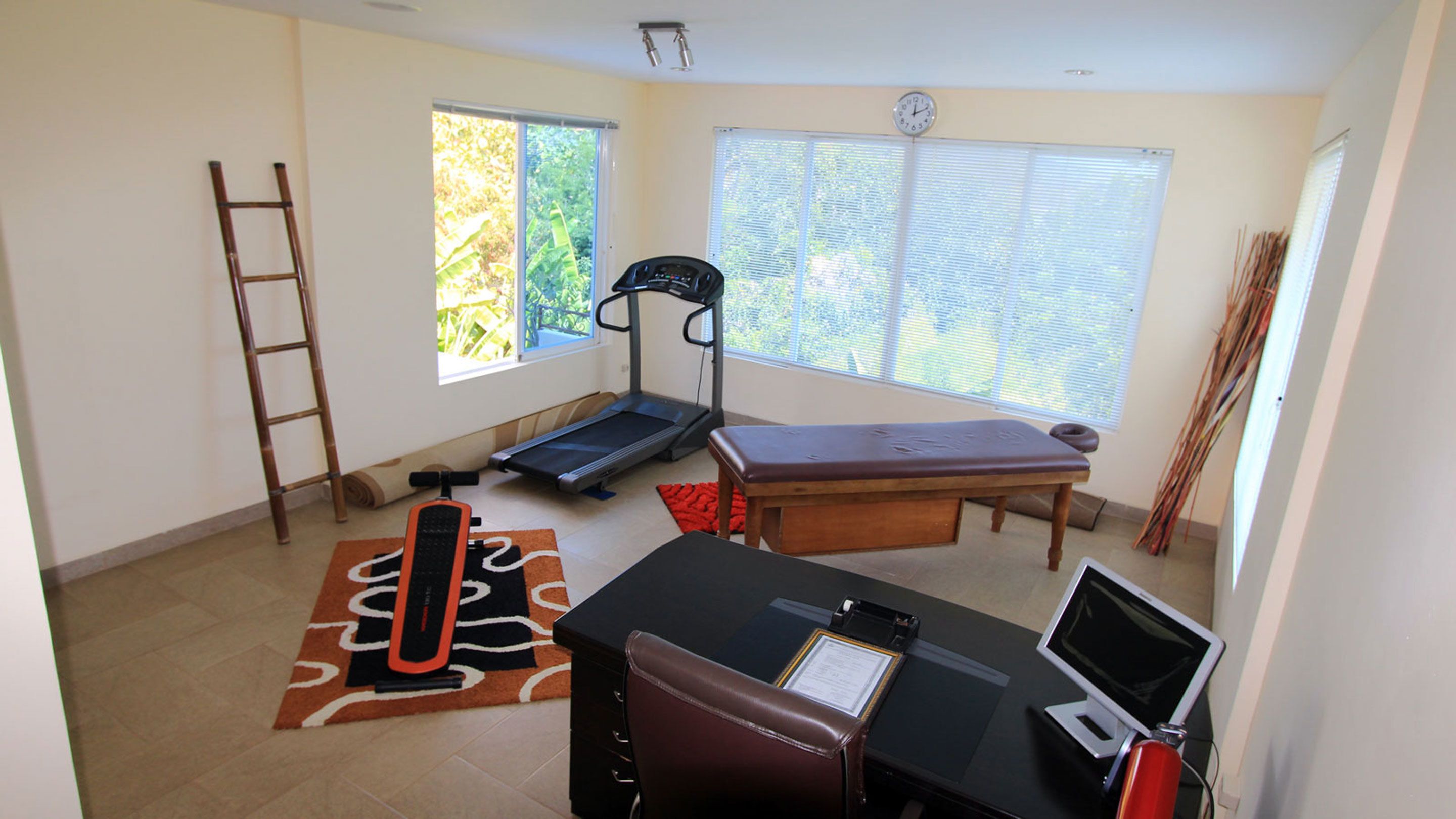 A room with a treadmill, a massage table, a desk with a computer, and a window with blinds.