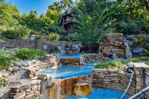A naturalistic water feature with cascading streams and a stone-built structure, surrounded by lush vegetation and a wooden building in the background.