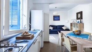 Une cuisine de taille réduite avec une fenêtre, un coin repas comprenant une table et des chaises, et un salon avec un canapé et une horloge au mur.