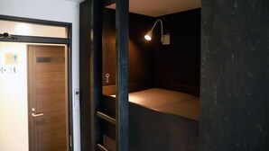 Una camera compatta con una porta in legno, un comodino, una lampada a muro e uno scaffale.