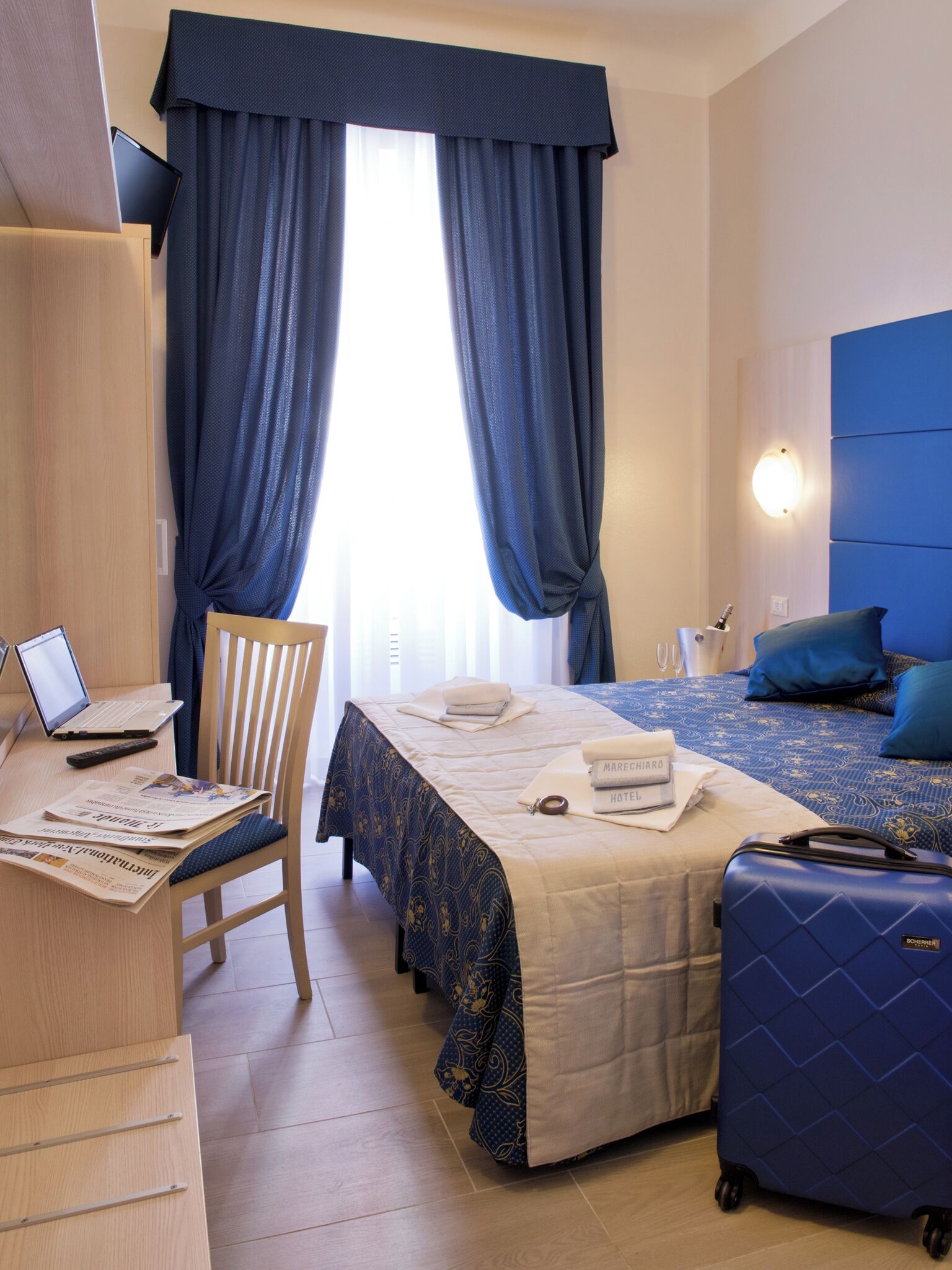 Una habitación de hotel con cama, escritorio con computadora portátil, silla, maleta y mesita de noche con lámpara.
