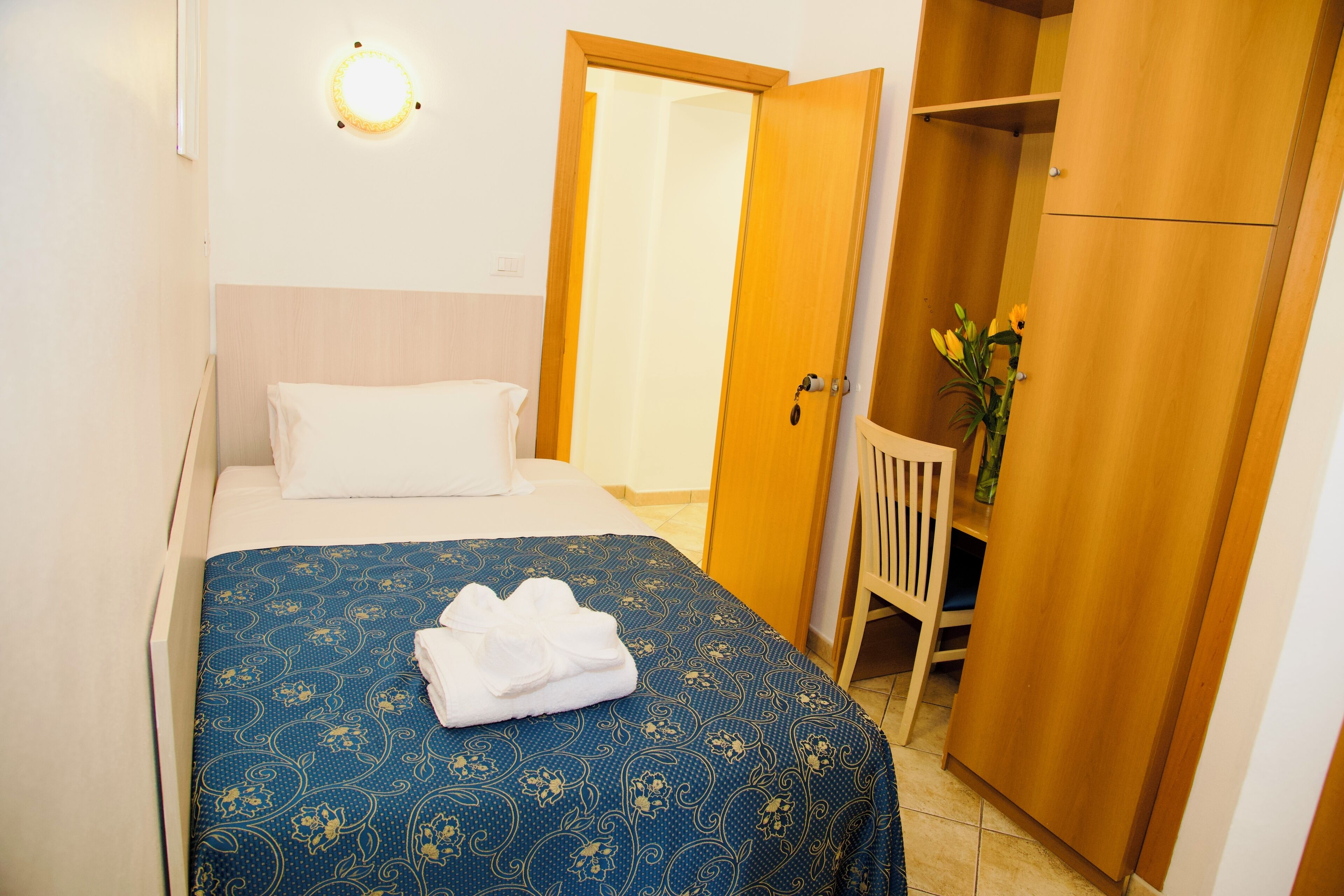 A neatly made bed with a blue patterned bedspread and white pillows, a wooden wardrobe, and a small table with a vase of flowers.