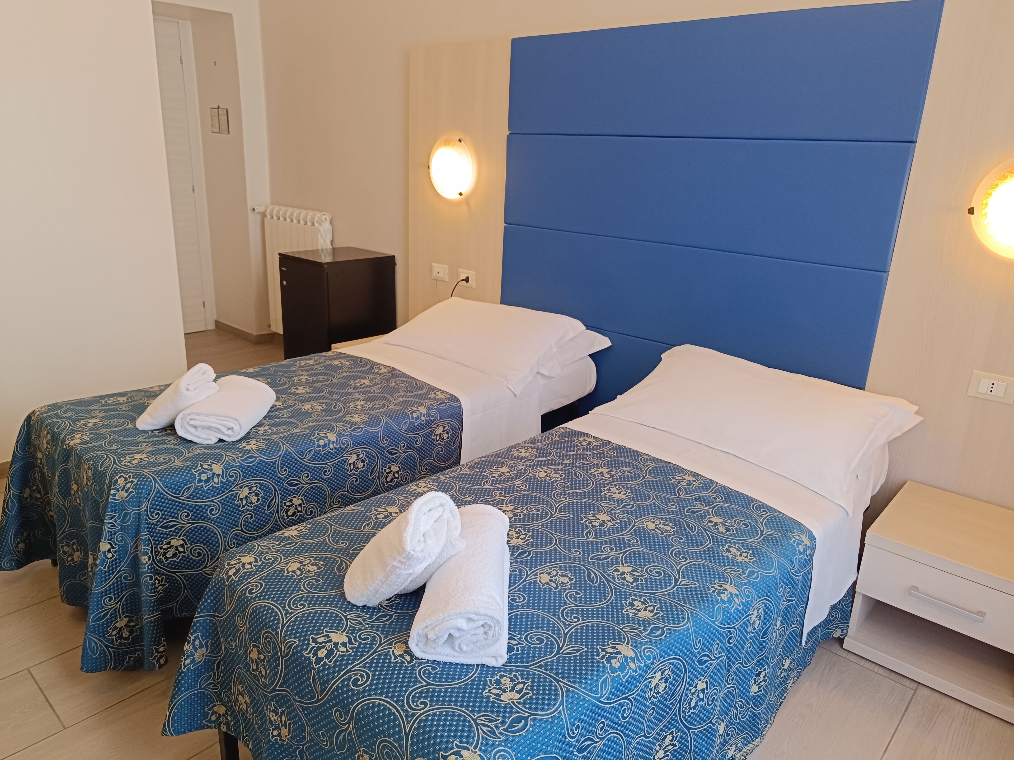 A hotel room with two beds, each with a blue patterned bedspread and white pillows. There are two wall-mounted lights, a small table, and a nightstand with a lamp.