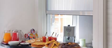 A breakfast buffet with various pastries, a coffee machine, and juice pitchers on a counter by a window.