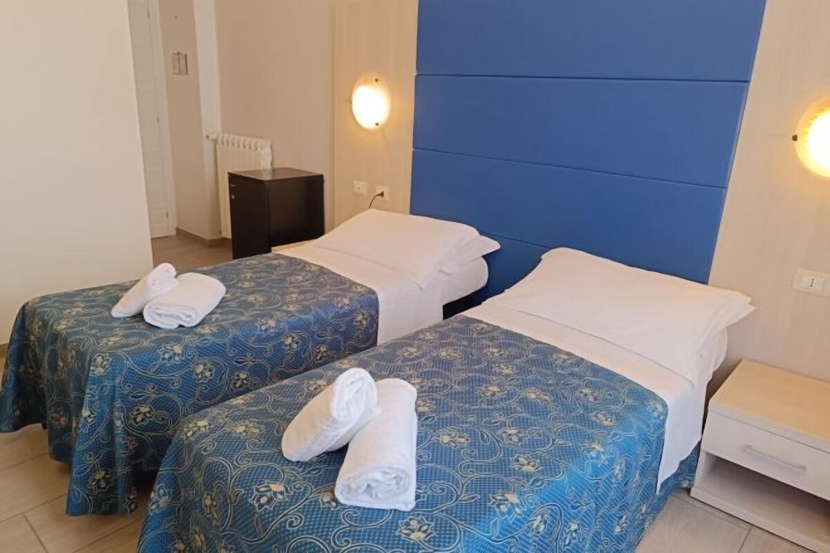 A hotel room with two beds, each with a blue patterned bedspread and white pillows. There are two wall-mounted lights on either side of the beds.