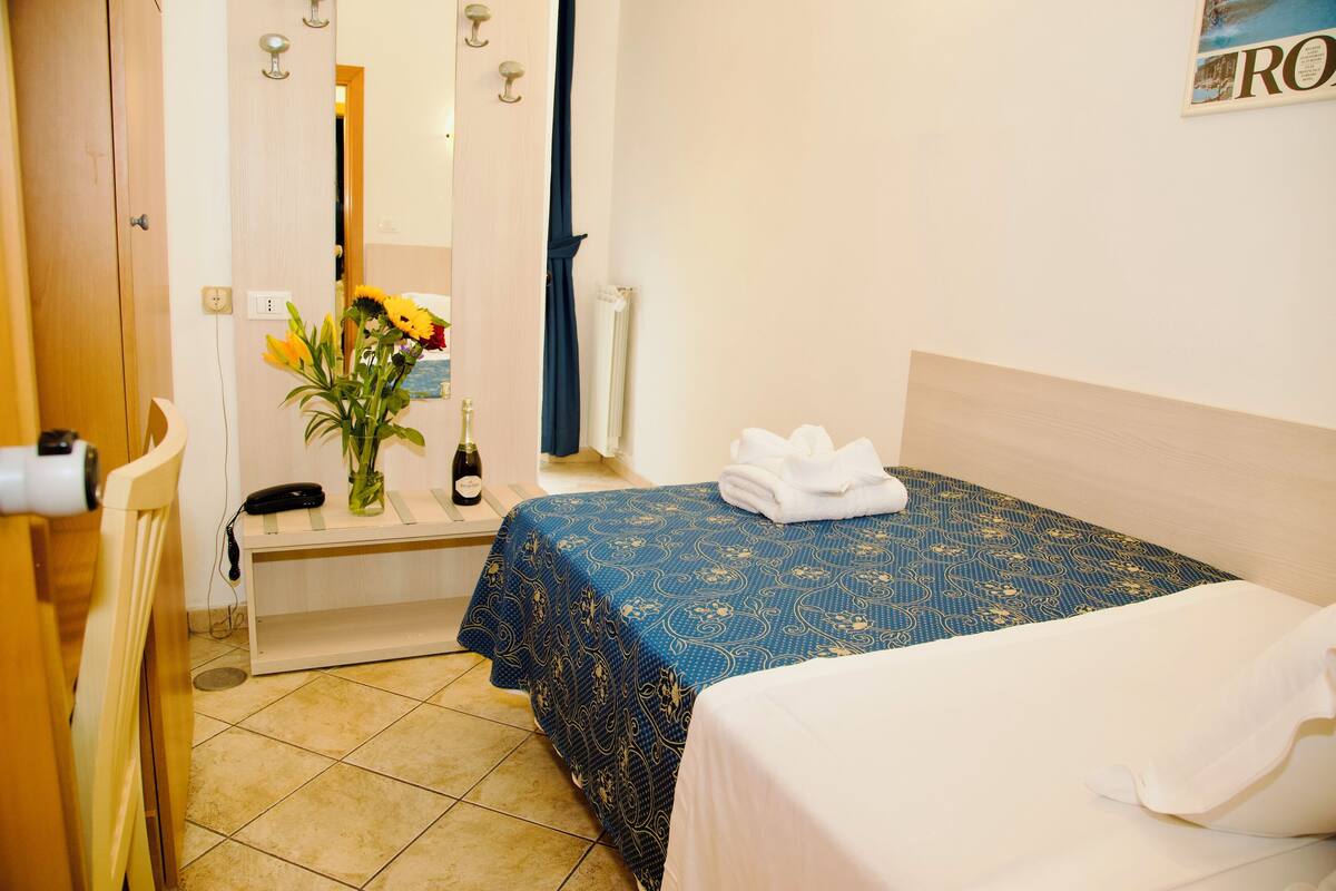 A hotel room with a bed, a desk with a vase of flowers, a bottle, and a chair.