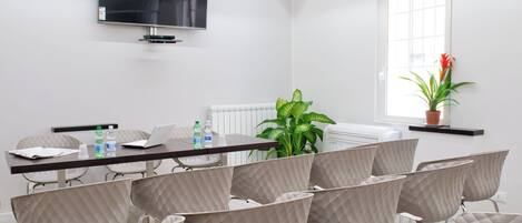 Sala de conferencias con una televisión de pantalla plana grande, un radiador, una planta en maceta y varias sillas acolchadas dispuestas alrededor de una mesa con botellas de agua y una computadora portátil.
