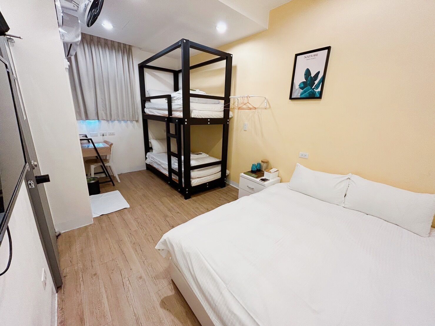 Una habitación de hotel con literas, un escritorio, una silla, una mesita de noche con lámpara y un cuadro enmarcado en la pared.