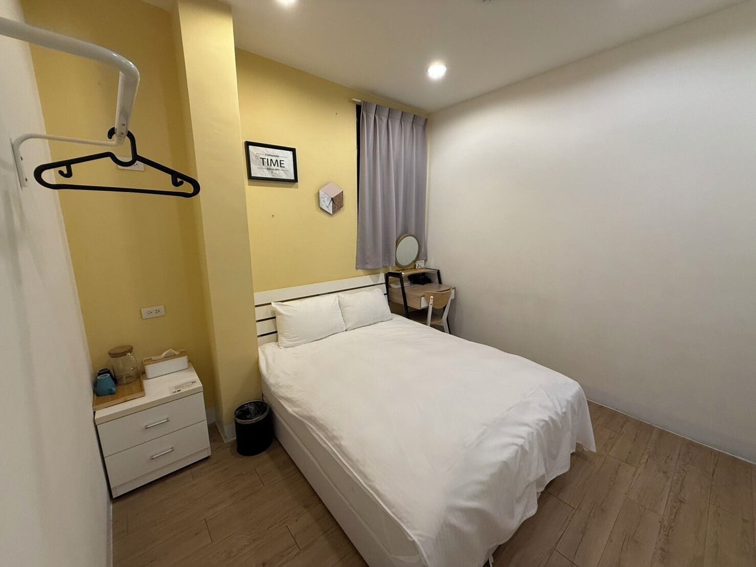 A bedroom with a bed, a bedside table, a mirror, a hanging light, and a wall-mounted clock.