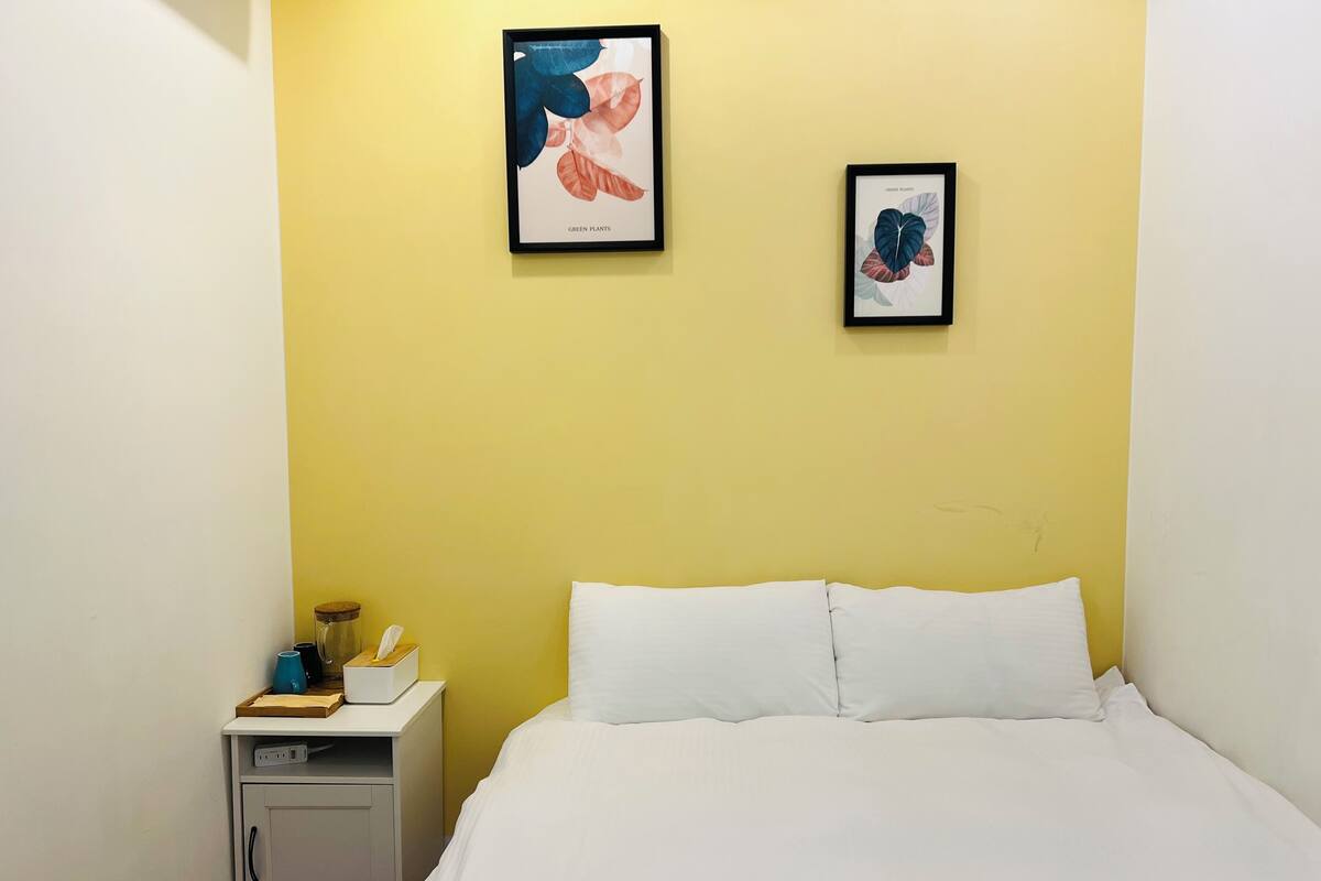 Quarto com cama, mesa de cabeceira e duas gravuras botânicas emolduradas na parede.