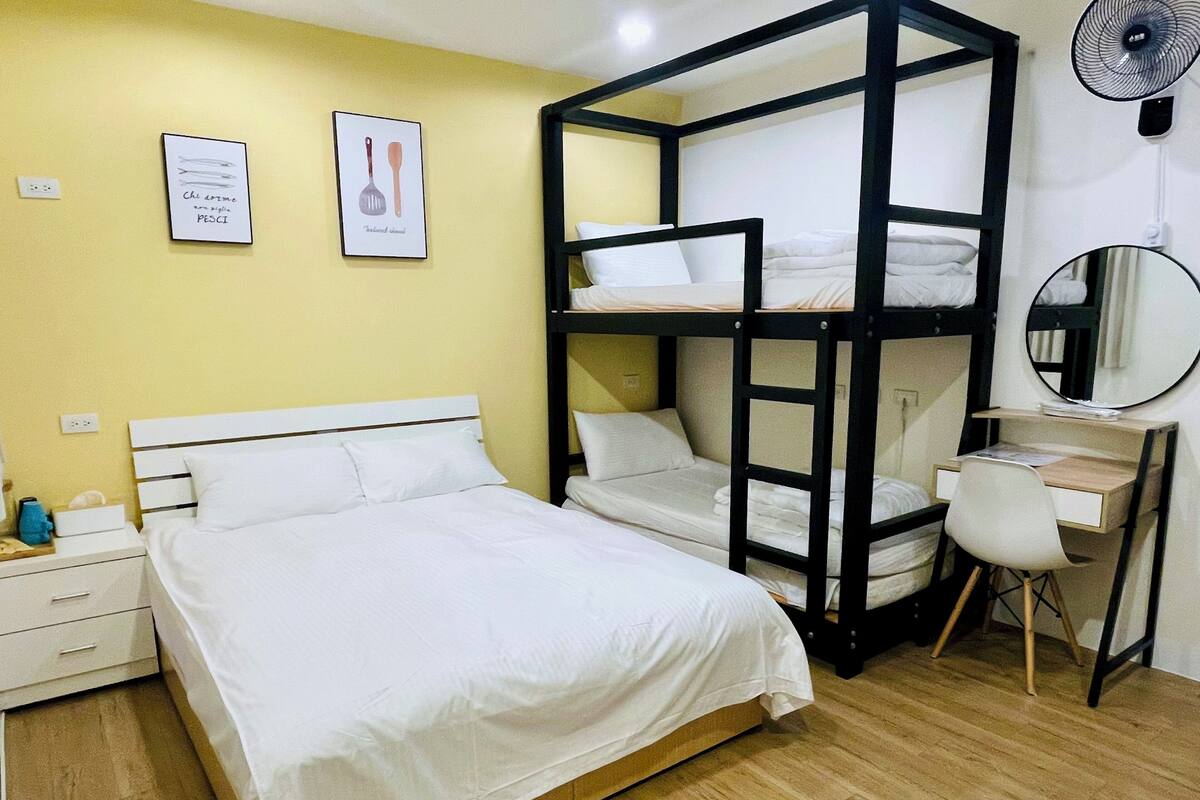 Quarto de hotel com uma cama beliche, uma escrivaninha com cadeira, um espelho e duas quadros na parede.