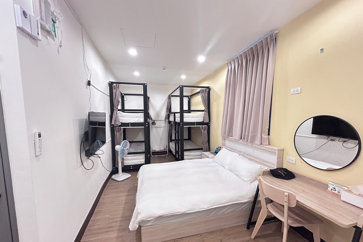 A hotel room with bunk beds, a desk, a chair, a bedside table, a mirror, and a wall-mounted fan.