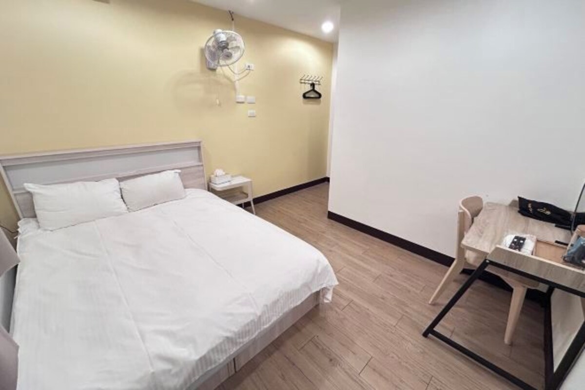 A hotel room with a bed, bedside table, desk, chair, and a wall-mounted fan.
