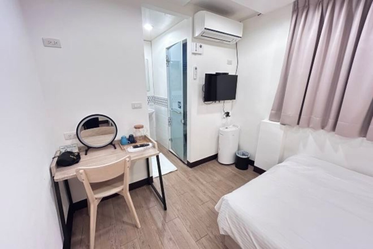 A hotel room with a bed, a desk with a chair, a mirror, a television, and a window with curtains.