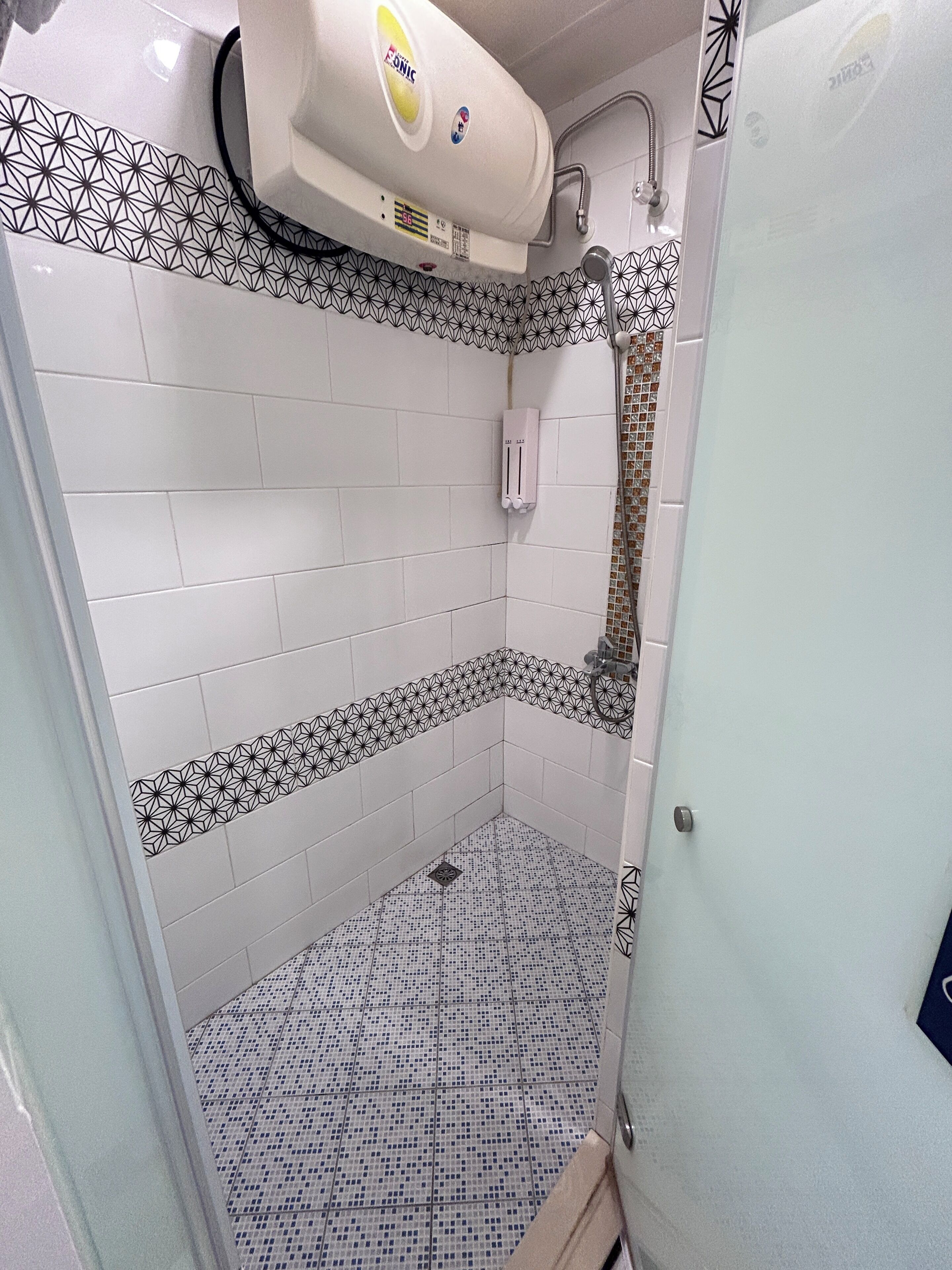 Casa de banho com paredes revestidas a azulejos brancos, chuveiro com porta de vidro e chão com padrão geométrico.