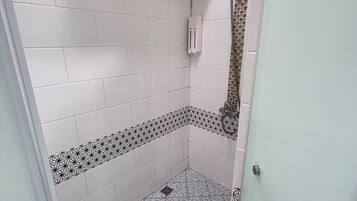 Casa de banho com paredes revestidas a azulejos brancos, chuveiro com porta de vidro e chão com padrão geométrico.