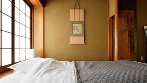 Una cama bien hecha con sábanas blancas y una manta gris, un cuadro en la pared, y un amplio ventanal que deja entrar luz natural.