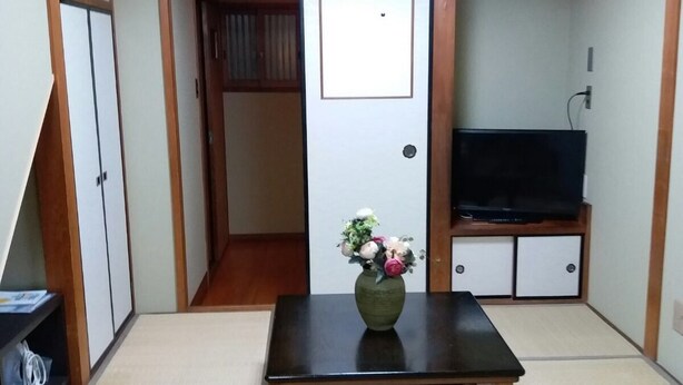 Una habitación japonesa tradicional con suelo de tatami, una mesa baja con un jarrón de flores, un televisor sobre un soporte de madera y puertas correderas.