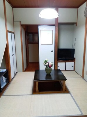 Een traditionele Japanse kamer met tatami vloeren, een lage tafel met een bloevas, een televisie op een houten standaard en schuifdeuren.