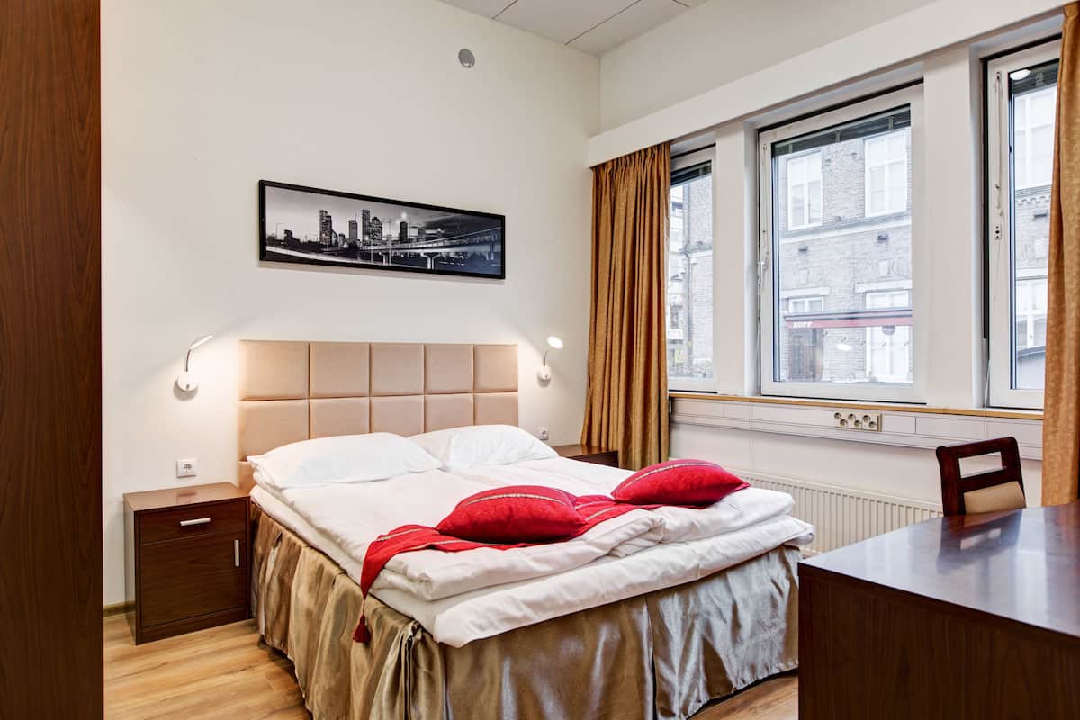 Habitación de hotel con cama, mesita de noche, escritorio, ventana con cortinas y una fotografía del paisaje urbano en la pared.